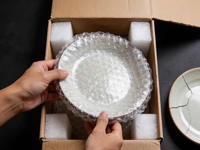 ceramic plate wrapped in bubble wrap inside cardboard box with foam corners showing fragile product packaging
